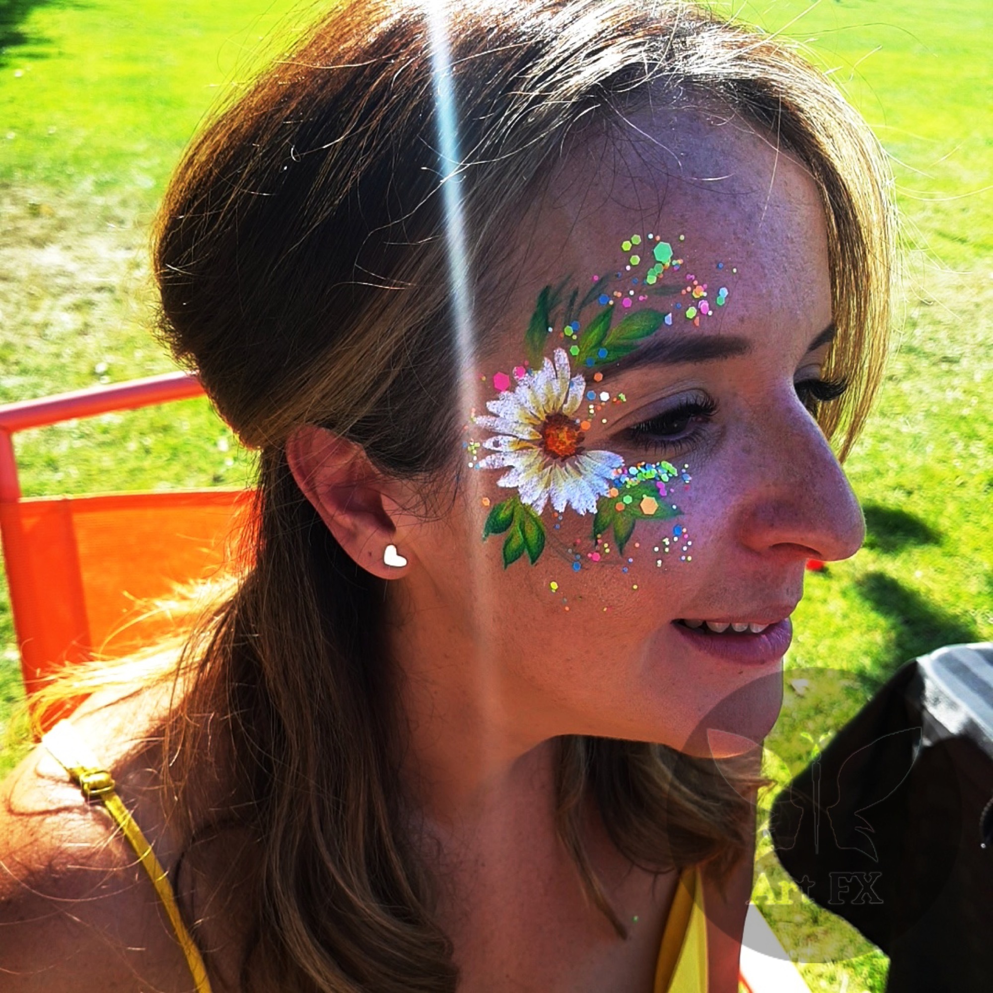 A woman with a vibrant tropical floral face paint design in pink, yellow, and green with chunky glitter.