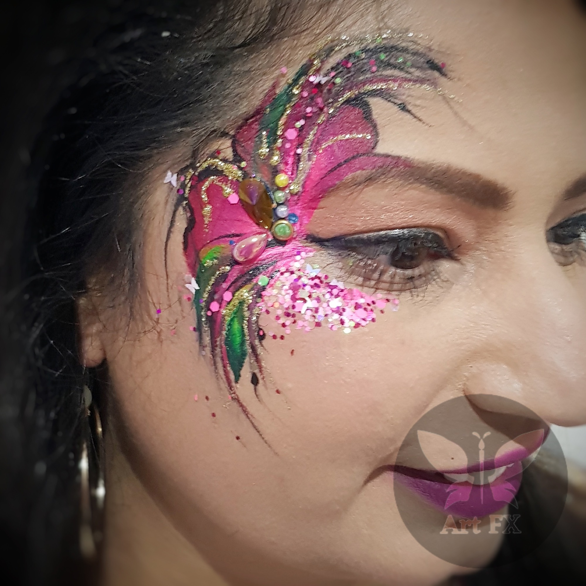 A woman carrying an intricate green and gold floral face paint design embellished with green and gold glitter.