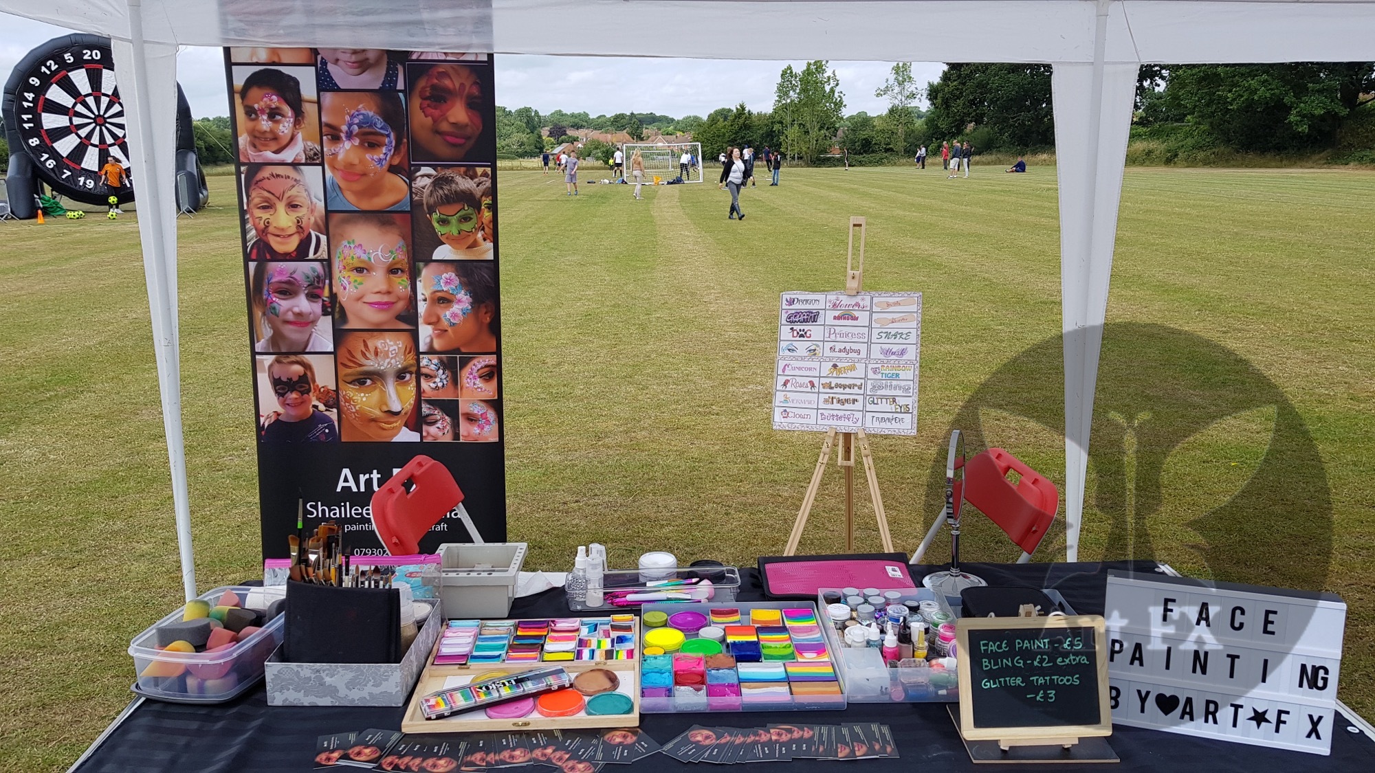 Professional face painting setup at an outdoor event with a display banner of colorful designs and a table of palettes.