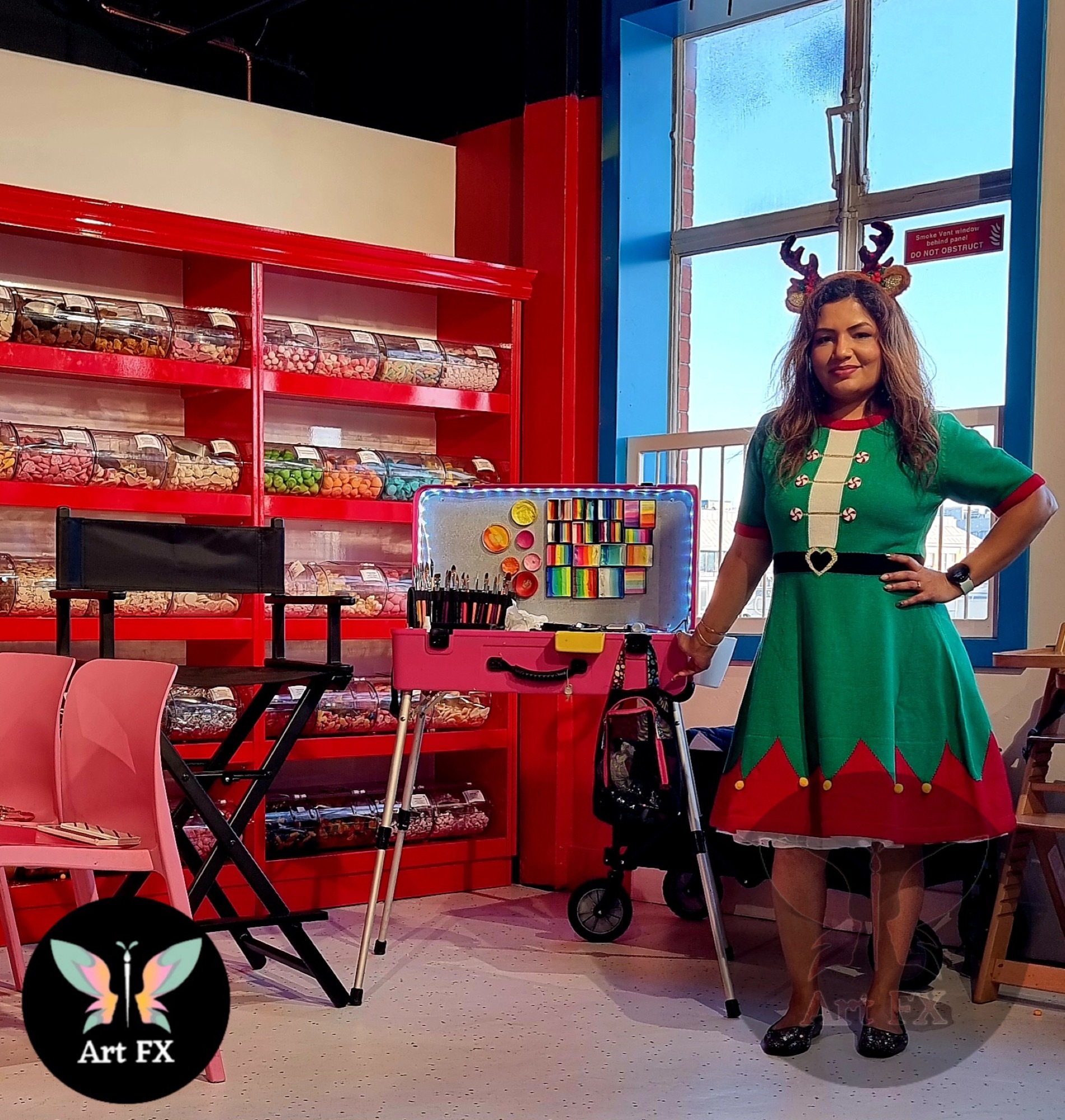 Shailee dressed in a festive elf costume standing next to her professional face painting kit in a colorful indoor setting.