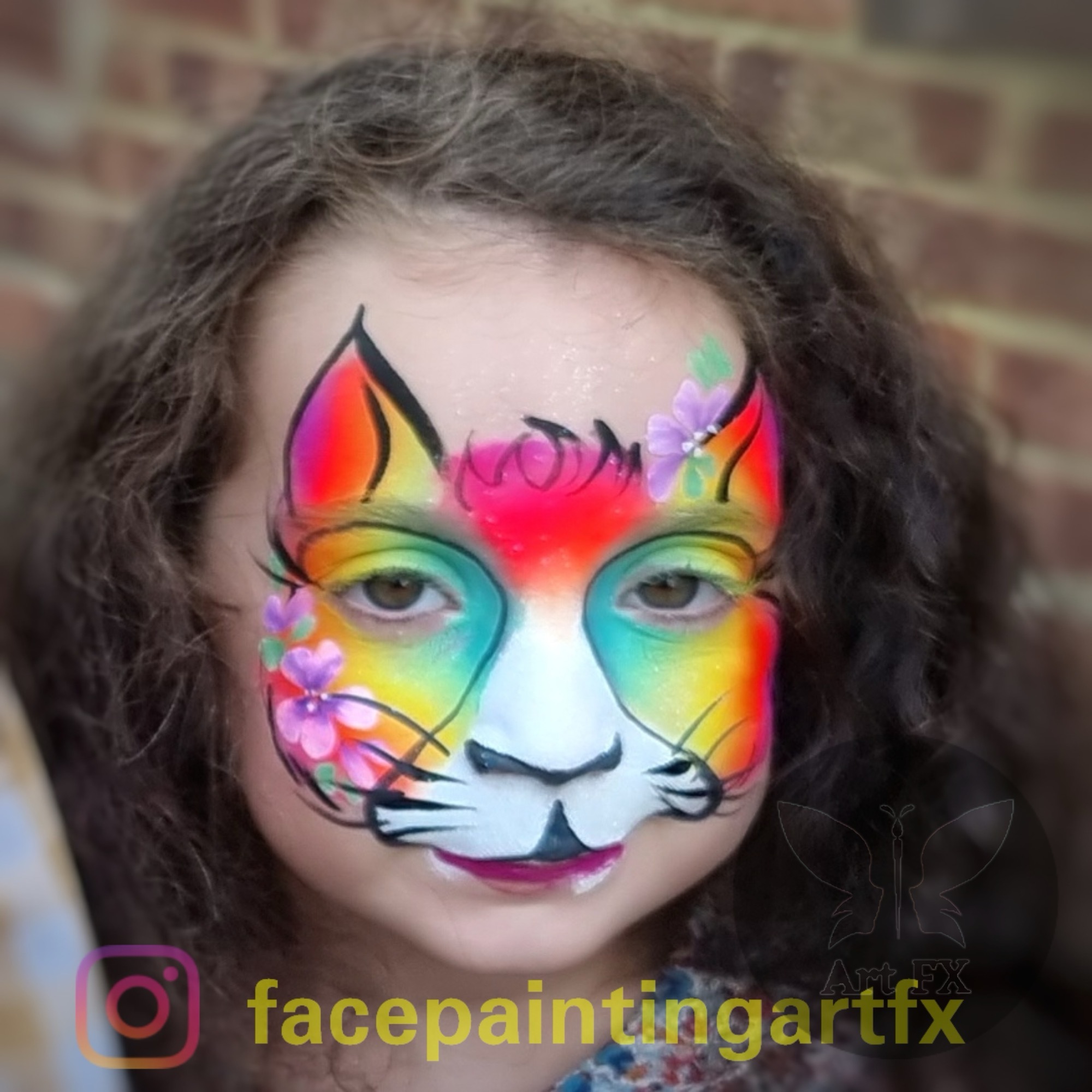 Young girl featuring a full-face rainbow cat face painting design with pink ears, a white muzzle, and flower details