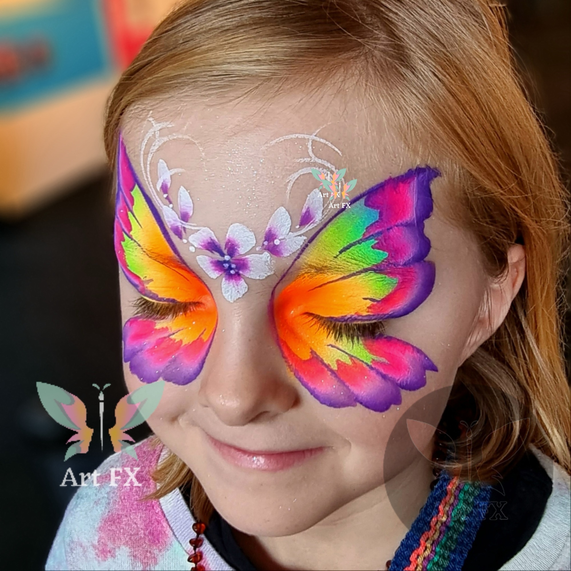 Young girl with eyes closed showcasing a bold rainbow butterfly mask face painting design with delicate white floral and swirl accents